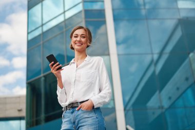 Modern ofis binasına karşı ayakta dururken akıllı akıllı telefonlu, gülümseyen genç bir iş kadınından. Şehirdeki cep telefonuyla mesajlaşan kendinden emin girişimci.