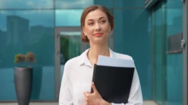 Ofis binasının dışında yürürken elinde klasör tutan kendine güvenen bir iş kadını. Akıllı giyinen genç ve mutlu bayan profesyonel dışarıda kameraya bakıyor.. 