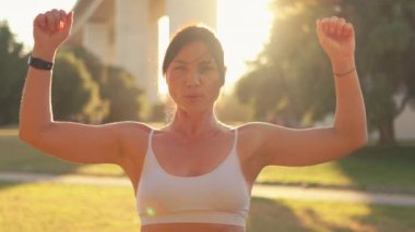 Parlak güneşli bir günde şehir parkında kollarını açan kararlı, sportif bir kadın. Spor giyimli genç bir kadın gündüz spor yaparken kameraya bakıyor.. 