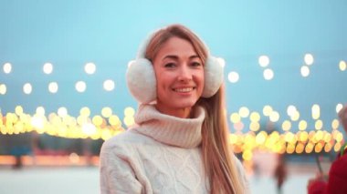  Kış zamanı. Noel tatili akşamı. Buz pateni pistinin dışında duran ve kameraya gülümseyen kulaklı bir kadın. Noel aydınlatması ve dekorasyon geçmişi. Kış tatili.