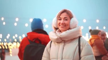 Kış zamanı. Noel tatili akşamı, kulaklıklı ve ceketli bir kadın buz pateni pistinin dışında kameraya bakıyor ve gülümsüyor. Noel aydınlatma ve dekorasyon arka planı.
