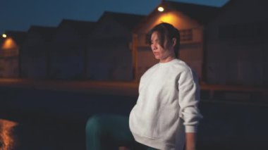 Sportif kadın geceleri şehirde gezinti güvertesinde çömelme ve bacak kaldırma egzersizleri yapıyor. Arka planda aydınlatılmış binalarla egzersiz yapan kararlı genç ve zinde kadınlardan oluşan bir vücut.. 