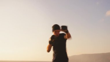 Gün batımında açık havada şapka antrenmanında aktif sporcu, elleriyle havada vuruyor. Kararlı eldivenli erkek sporcu gün batımında gökyüzünün altında boks yapıyor..