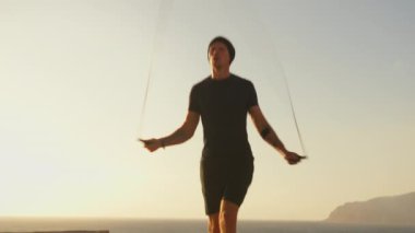 İp atlayan ve idman, atlama ya da antrenman için dışarıda egzersiz yapan bir adam. Sporcu gün batımında kardiyo, fitness ve enerji için denize karşı vücut sağlığı ve sağlık motivasyonu