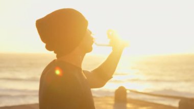 Sportif sporcu gün batımında gökyüzü altında şişeden denize su içiyor. Yoğun egzersizden sonra mola veren şapkalı genç bir erkek koşucu. Sağlıklı yaşam tarzı.