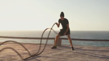 Atletik erkek fitness model eğitim ipleri gün batımında denize karşı savaşıyor. Spor fitness sağlıklı yaşam tarzı konsepti. Beyaz tenli, kaslı vücut fonksiyonel eğitim dayanıklılığı.