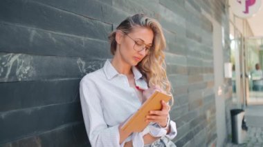 Gözlüklü düşünceli, yaratıcı bir kadın gelecekteki iş planlarını düşünüyor, not defterine notlar alıyor yaratıcı fikirler yazıyor ve gülümsüyor. Karanlık duvarın yanında duruyor..