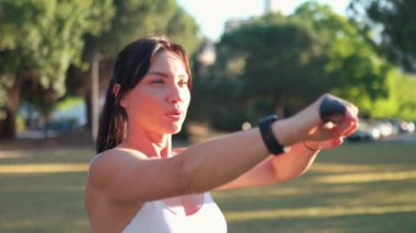 Parlak güneşli bir günde şehir parkında direniş bandıyla egzersiz yapan kararlı sporcu bir kadın. Aktif giyimli genç bir kadın çimenlikte kol egzersizi yapıyor..