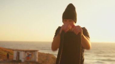 Gün batımında deniz kıyısındaki plajda boks yapan bir sporcu. Kararlı erkek savaş halatlarını tutuyor elleriyle vuruyor, antrenman yapıyor, gün batımında spor yapıyor. Formda, sporcu, sağlıklı yaşam tarzı..