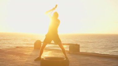 Atletin tekere çekiçle vurması. Güç ve kararlılık kavramı. Güneş batarken turuncu gökyüzü altında deniz kenarında antrenman yapan sporcu.