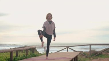 Orta yaşlı kadın yoga yapıyor. Etrafı okyanus dalgaları ve bulutlu gökyüzü sesleri ile çevrili. Nazik hareketleri ve sakin ifadesi, uygulamasının meditasyon yönünü vurguluyor.