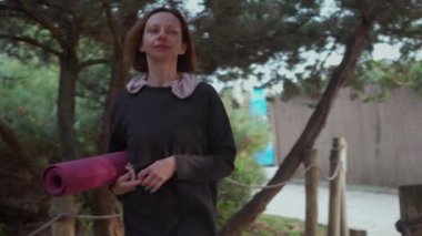 Kadın elinde yoga minderiyle yemyeşil parkta yürüyor. Huzurlu bir ortam hazırlıyor ve yoga deneyimini canlandırıyor.