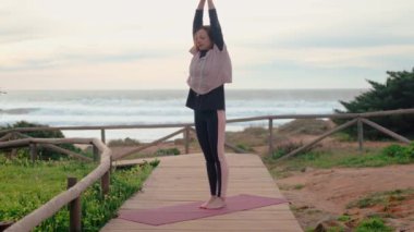 Orta yaşlı bir kadın, okyanus dalgalarının sesi ve bulutlu gökyüzü eşliğinde sakin plajda yoga yapıyor. Sakin hareketleri huzuru ve farkındalığı yansıtıyor, sağlık rutini için mükemmel.