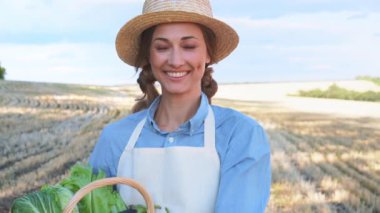 Çiftlik arazisinde sepet dolusu sebze tutan mutlu kadın çiftçi. Güneşli bir günde önlük ve hasır şapka takan kendinden emin kadın tarım uzmanına yakınlaş.. 