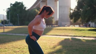 Kararlı sportif kadın güneşli bir günde çimlere atlıyor. Şehir parkında ısınan spor giysili genç bir kadının yan görüntüsü.. 