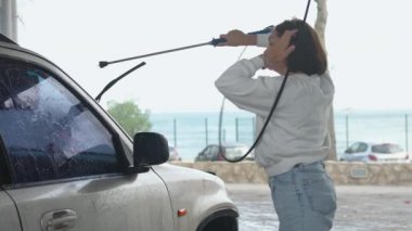 Otomobil yıkama istasyonunda araba yıkayan genç bir kadın. Gündelik elbiseli bir kadın şehirde basınçlı su dolu bir hortum kullanıyor.. 