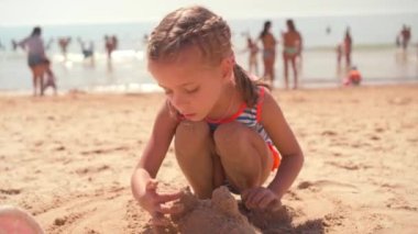Çizgili mayo giymiş küçük bir kız okyanus kıyısında kumdan kale inşa ediyor. Deniz kenarında çocukluğun neşeli ve masum anlarını yakalar.