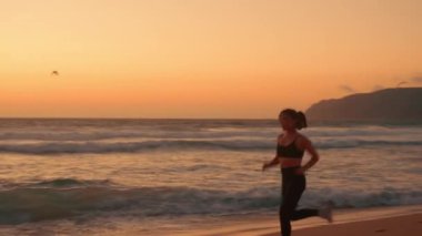 Gün batımında sahilde koşan kararlı bayan koşucu. Heyecanlı sporcu kadın kumlu sahildeki koşusunun sonunda havada ikiye bölünüyor.. 