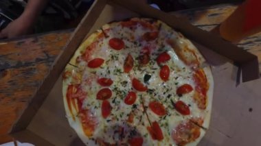 Yukarıdan bakıldığında, bir akşam panayırında birkaç kişi pizza dilimleri için uzanır. Sadece elleri görünür, canlı ve ortak bir atmosfer yaratır.