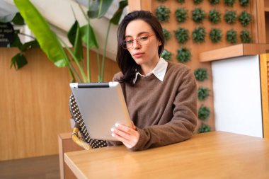 Ofis masasında oturmuş dijital tablet kullanırken gözlüklü bir kadın ekranı seyrediyor. Tableti elinde tutan kadın sosyal medyada geziniyor