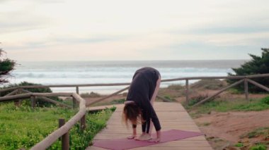 Orta yaşlı kadın yoga yapıyor. Arkaplanı okyanus dalgaları ve bulutlu gökyüzü. Hareketleri zarif ve odaklanmış. Huzurlu ve meditasyon rutinini gösteriyor.