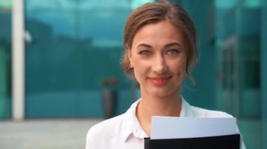 Ofis binasının dışında yürürken elinde klasör tutan kendine güvenen mutlu iş kadını. Güzel genç bayan profesyonel gündüz vakti gülümsüyor ve kameraya bakıyor.. 