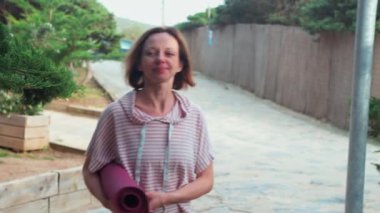 Kadın yoga meraklısı, elinde minderiyle resim parkında geziniyor ve huzur verici ve canlandırıcı yoga pratiğini kucaklamaya hazır.