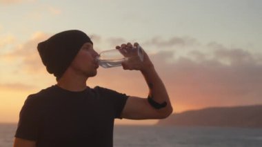 Sporcu gün batımında deniz kenarında antrenmandan sonra plastik şişeden su içer. Mutlu, genç, yakışıklı, kasketli beyaz adam doğada koştuktan sonra dinleniyor, soğuk su içiyor..