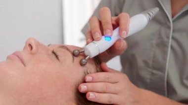 Kozmetik uzmanı müşterilerin yüzünde iyontoforez kullanıyor. Kadın eli, kadın yüzü boyunca hareket eden top kafalı makine. Beyaz kadın yüz derisi için fizyoterapi görüyor.. 