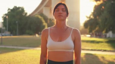Sportif bir kadın parkta meditasyon yapıyor. Kamera, kadın yüzüne doğru yakınlaşır gözlerini açar ve kameraya bakar. Spor sutyenli kadın sağlıklı yaşam tarzını temsil ediyor..