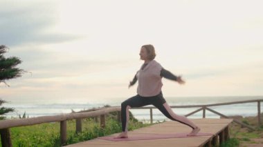 Orta yaşlı bir kadının bulutlu arka planla yoga çalışmasını izleyin. Vdeo onun meditasyon hareketlerini ve okyanusun sakin atmosferini yakalar. Vücut ve doğa arasındaki uyumu ön plana çıkarır.