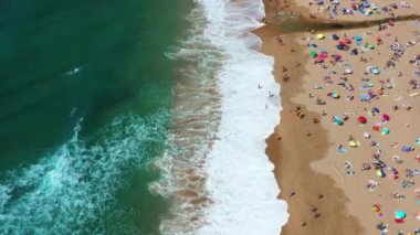 Magoito Plajı 'ndaki kumlu sahil şeridinde isimsiz insanların ve şemsiyelerin insansız hava aracı görüntüsü. Portekiz 'in Sintra kentindeki ünlü sahil şeridine yaz boyunca deniz dalgaları çarpıyor.. 