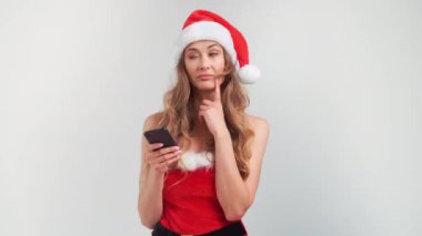 Kafası karışmış Noel Baba kadın gri arka planda dururken akıllı telefon kullanıyor. Sade stüdyoda alet kullanan düşünceli bir kadının portresi.. 