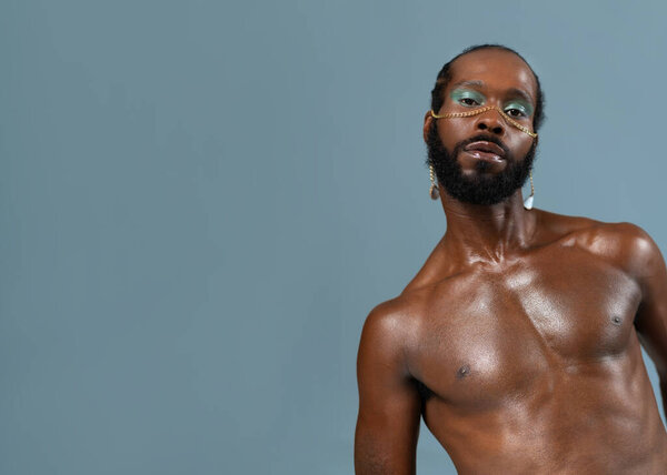 Bearded African American gay man with light makeup and gold accessory on face. Shirtless adult homosexual male looking at camera with mockup space on blue background.