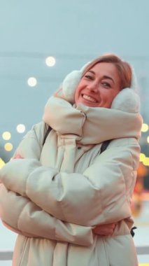 Sıcak beyaz ceketli ve kürklü bir kadın Noel ışıklarıyla çevrili bir buz pateni pistinin yanında duruyor ve kameraya gülümsüyor, neşeli tatil atmosferini kucaklarken mutluluk saçıyor.