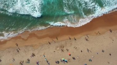 Yukarıdan bakıldığında, kumlu sahildeki meçhul insanların hafif dalgalar kıyıya ulaştığı görülüyor. Dalgalar ritmik desenler oluşturur, canlı ama sakin plaj sahnesine eklenir. 