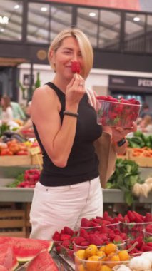 Orta yaşlı bir kadın pazarda çilek seçiyor, meyveleri kokluyor, sağlıklı beslenmeye odaklanıyor. 