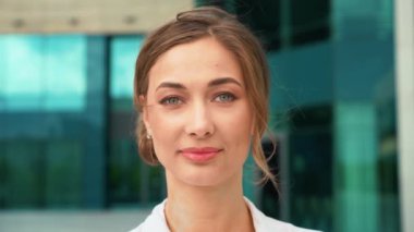 Doğrudan kameraya bakan kendine güvenen bir iş kadınının yakın plan portresi. Modern ofis binalarının önünde duruyor, şehir ortamında profesyonelliği ve başarıyı somutlaştırıyor.