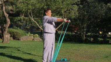 Aktif yaşlı bir kadın parkta kollarını açmak için direniş bandı kullanıyor. Etrafı ağaçlar ve doğa ile çevrili. Sağlıklı ve zinde kalmaya kararlı olduğunu gösteriyor. 