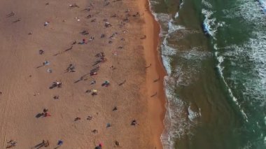 Okyanus dalgaları sıcak kuma çarparken turistlerin güneşlenip dinlendiği kalabalık bir plajın insansız hava aracı görüntüleri. 