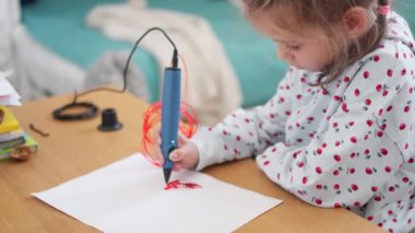 Küçük kız odasında masasında oturuyor. Plastik çizimler yapmak için 3 boyutlu kalem kullanıyor. Renkli plastikle şekiller ve tasarımlara odaklanıyor. 