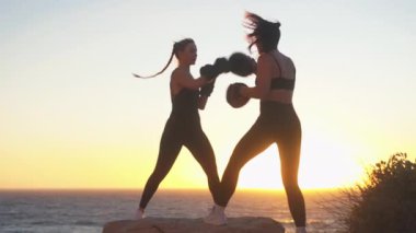 Parlak günbatımında deniz manzaralı kaya ve gökyüzünde boks antrenmanı yapan sportif kadın dövüşçüler. Kararlı genç bayan boksörler sahilde antrenman yapıyor.