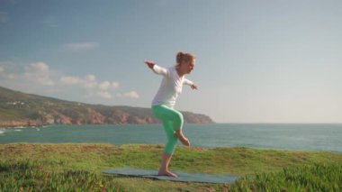 Son sınıf öğrencisi kadın, poz vermek için okyanus kenarında yoga yapıyor. Bir amatör olarak zorluklarla karşılaşır ama denge ve sükuneti bulma azmi sakin kıyı ortamında parlar.