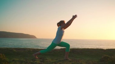 Yaşlı kadın, savaşçı pozuyla yoga yapıyor. Arkasında okyanus ve dağların ardında güneş batarken, vücudu zar zor görünüyor. Huzurlu bir siluet oluşturuyor.