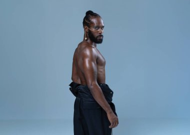 Ciddi bir Afro-Amerikan tişörtsüz gay model gri arka planda duruyor. Stüdyoda belinde ceket olan kibirli homoseksüel bir adamın portresi.. 