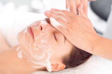 Profesyonel kozmetoloji: Kozmetik salonunda kadınların yüzüne krem süren ve masaj yapan terapistlerin elleri. Rahatlamış bayan müşteri, profesyonel olarak kaplıcada şımartılıyor.. 