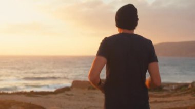 Güneş batarken okyanus sahilinde koşan atletik bir adam. Kafkasyalı erkek, turuncu gökyüzünün altında deniz sahilinde koşu antrenmanı yapıyor..