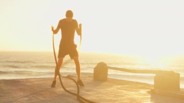 Kararlı sportif adam gün batımında açık gökyüzüne karşı savaş halatlarıyla havada boks yapıyor. Genç ve formda bir erkek, doğada çapraz uyum eğitimi alıyor.. 