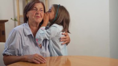 Neşeli torun, gülen büyükannesinin kulağına evde bir şeyler fısıldıyor. Samimi ortam, sevgi ve mutlulukla dolu sıcak, sevgi dolu bir anı yakalıyor.