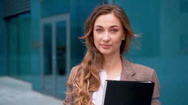 İş kadını, uzun saçlı, kameraya gülümseyen beyaz gömlekli, kahverengi ceketli, ofis binalarının yanında duran profesyonel ve başarılı bir iş imajı sergileyen bir iş kadını.